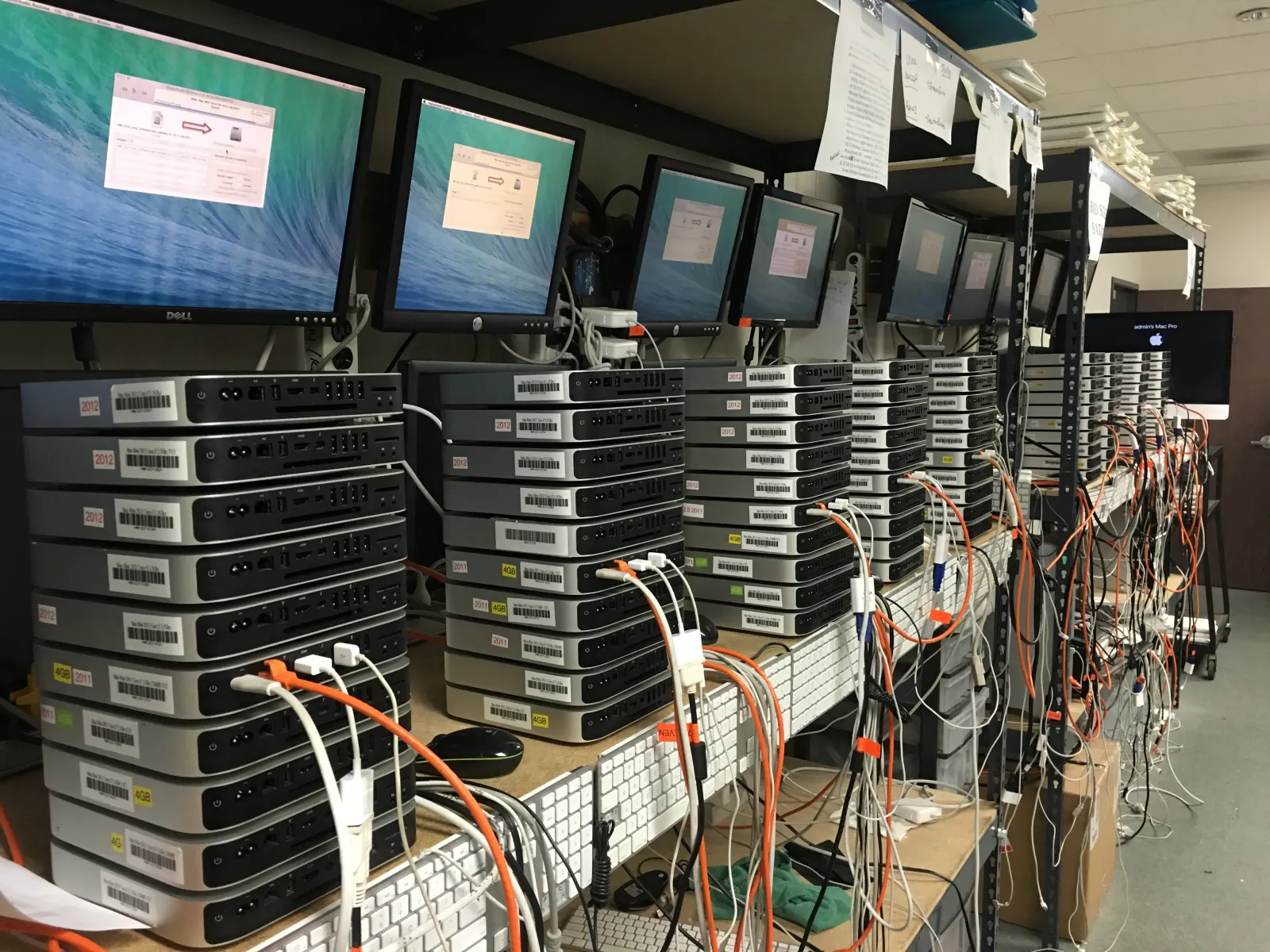 Stacks of Mac minis on a bench, cabled to the network with monitors above, being set up before they go out