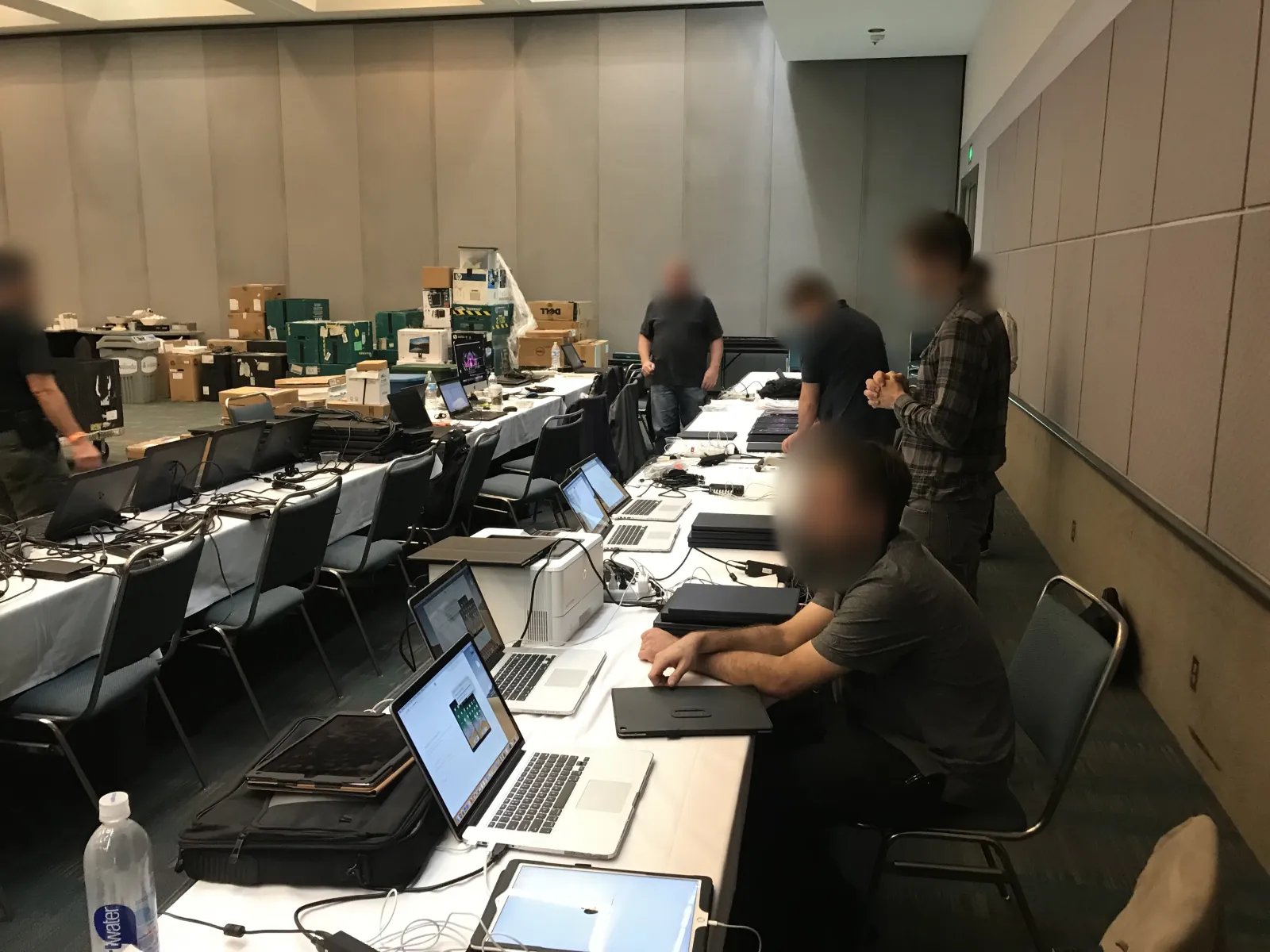 Mac Rentals staff setting up and testing laptops on long tables in a meeting hall before an event, faces blurred