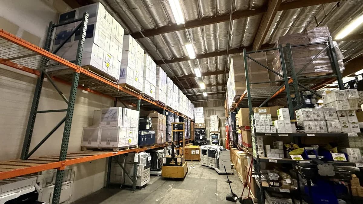 Inside the Mac Rentals warehouse: pallet racking stacked with boxed equipment, copiers on the floor and a pallet jack in the aisle