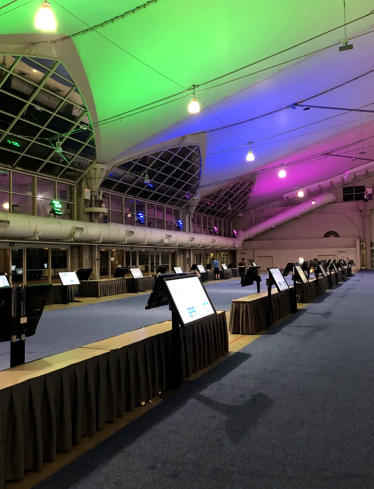 Check-in screens on floor stands at the edge of a convention hall lit in purple and green